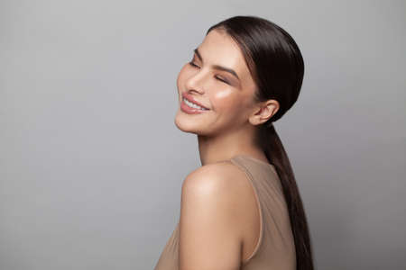 Pretty woman with closed eyes and dark eyebrows smiling on gray background. Attractive female model with light nude make-up, beauty photo, copy space, close upの写真素材