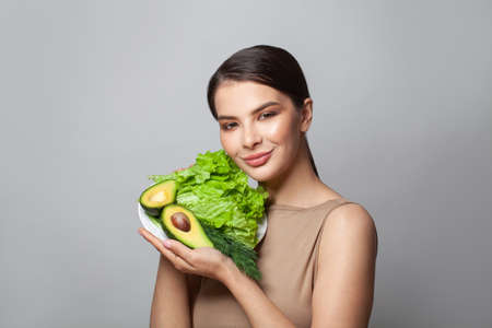 Portrait of young woman showing avocado and green lettuce on gray background