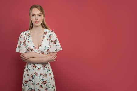 Pretty happy female with ginger hair, dressed casually, looking with satisfaction at camera portrait. Studio shot of beautiful woman standing against red studio wall.の写真素材