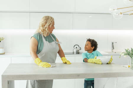 Caring mom teaching her son how to clean table with detergent in white kitchenの写真素材