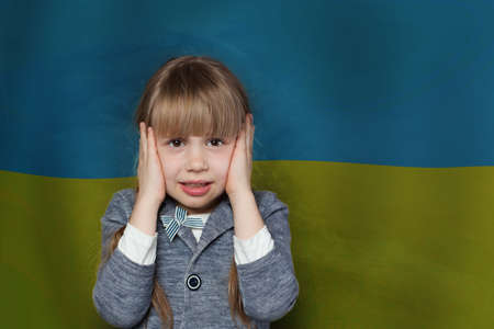 Frightened child girl covers her ears with her hands so as not to hear anything against Ukrainian flag background. Stop war in Ukraineの写真素材