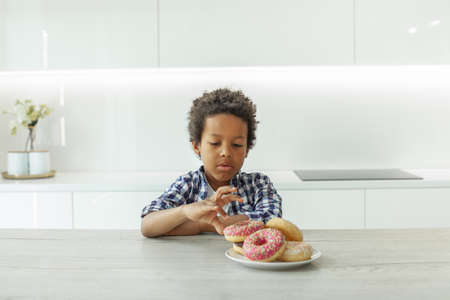 Cute kid boy with black hair eating donuts in white kitchenの写真素材