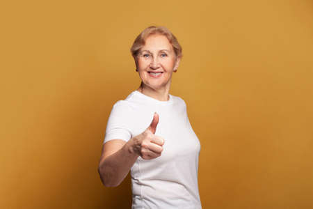Happy senior woman in white t-shirt showing thumb up on bright yellow studio wall banner backgroundの写真素材