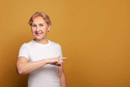 Senior woman in white t-shirt smiling and pointing finger at copy space on bright yellow studio wall banner backgroundの写真素材