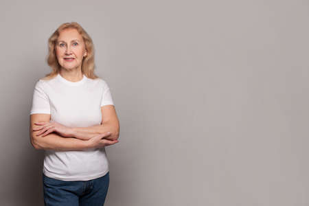 Optimistic confident senior woman wearing white t-shirt standing with crossed arms on gray studio wall backgroundの写真素材