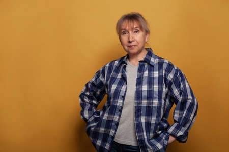 Cute smiling senior woman in blue shirt standing against vivid yellow studio wall backgroundの写真素材