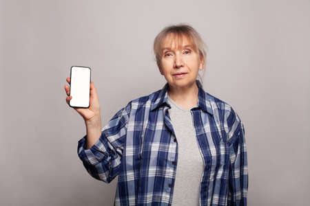 Happy senior woman showing modern smartphone with white empty display on grey studio wall background...