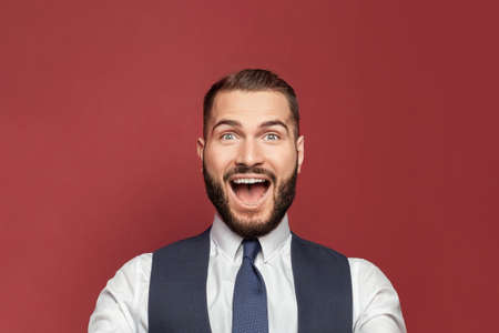 Young man with a surprised happy expression won on red studio background. Human facial emotions and...