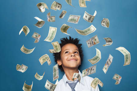 Successful little black kid boy standing under us dollar money rain on blue backgroundの写真素材