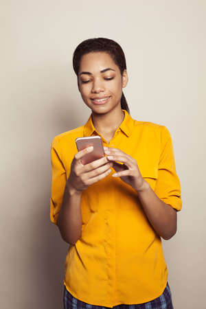 Cheerful black woman wearing casual yellow shirt holding smartphone against white studio wall backgroundの写真素材