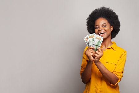 Happy young woman holding hundreds of dollar money banknotes and celebrating success on grey background. Finance, currency and people conceptの写真素材