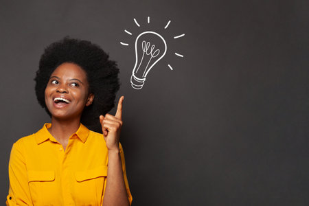 Beautiful smiling african american woman have an idea and pointing up to light bulb on black backgroundの写真素材