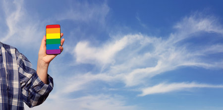Female hand holding smartphone with LGBT glag on the background of cloudy sky. Support, stop discrimination conceptの写真素材