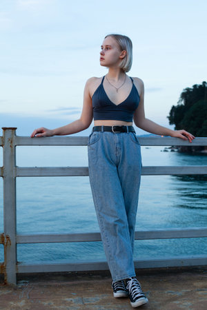Pretty woman in jeans and tank top standing on pier on the background of blue sea and light cloudy...