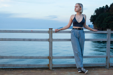 Young woman in jeans and blue tank top standing on pier on the background of blue sea and light...