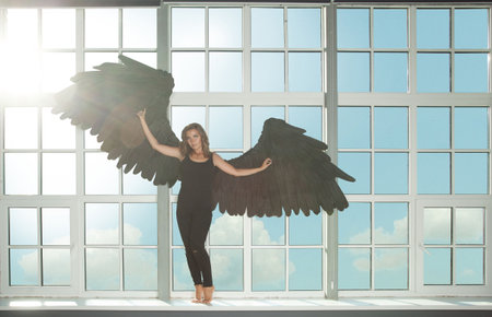 Beautiful perfect woman dark angel against window and sky with clouds background