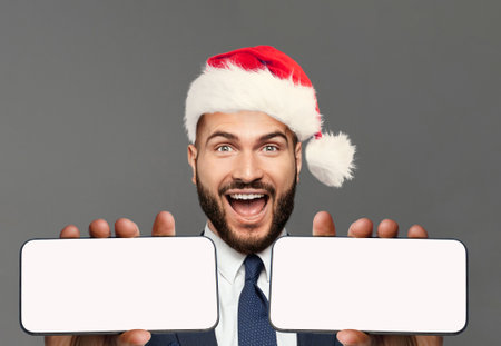 Happy excited businessman in Santa hat showing two smart phones. Smartphone with blank empty white screen display for new Application or mobile website, Mockup bannerの写真素材