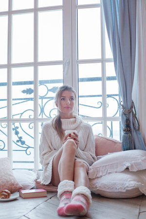 Pretty smiling lady in white sweater holding hot drink, resting, sitting on wooden floor in front of the window at homeの写真素材