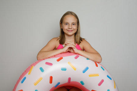 Smiling child girl with pink rubber inflatable ring swimming pool float standing on white studio...
