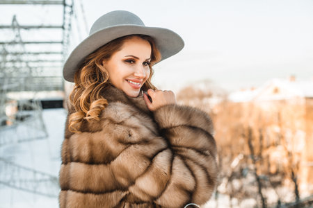 Portrait of smiling brunette woman wearing fur winter coat and gray hat on the background of cityの写真素材