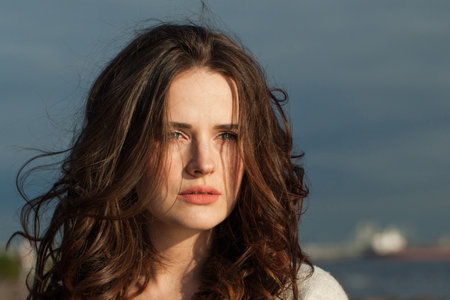 Attractive brunette woman with fresh skin, natural make-up and healthy wavy brown hair enjoying the sunny morning and sea air. Female model outdoor fashion portraitの写真素材
