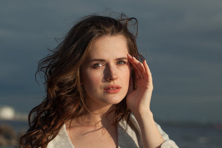 Thoughtful brunette woman with fresh skin, natural make-up and healthy wavy brown hair enjoying the sunny morning and sea air. Female model outdoor fashion portraitの写真素材