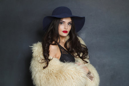 Glorious fashion model woman with long dark wavy hairstyle and red lips make-up wearing fashionable hat beauty studio portraitの写真素材