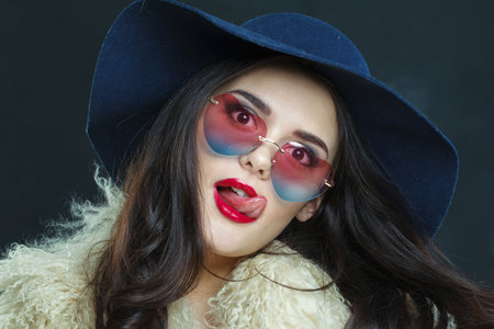 Funny fashion model woman with long dark wavy hairstyle and red lips make-up wearing fashionable hat sticking out your tongue beauty studio portraitの写真素材