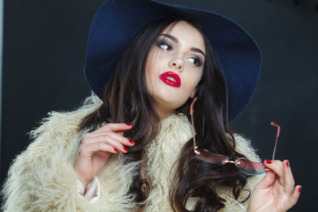 Glamorous fashion model woman with long dark wavy hairstyle and red lips make-up wearing fashionable hat beauty studio portraitの写真素材