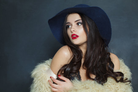 Sensual fashion model woman with long dark wavy hairstyle and red lips make-up wearing fashionable hat beauty studio portraitの写真素材