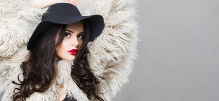 Dancing young adult woman fashion model with bright make-up and long wavy hair wearing blue hat posing against white studio wall backgroundの写真素材