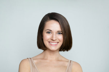 Positive young female model with healthy hair, makeup and shiny clear skin posing against gray studio wall background. Facial treatment, cosmetology, hair care and beauty woman conceptの写真素材