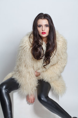 Elegant sensual brunette woman with long hair wearing black leather pants, high heels and a white fluffy coat posing against white studio wall backgroundの写真素材