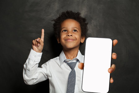 Joyful little school boy holding cell phone in his hand presenting smartphone empty screen display on blue studio wall background. Smartphone display mockupの写真素材