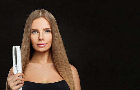 Young smiling woman with long straight healthy hairstyle using straightening iron on black background. Beauty, hair care, hair treatment and cosmetic conceptの写真素材