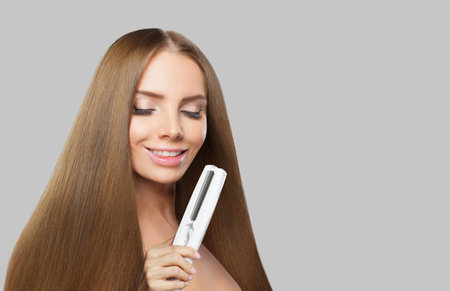 Cheerful brunette lady with straight hair holding straightening iron on white background. Female beauty, coiffure, hair care, cosmetic and styling conceptの写真素材