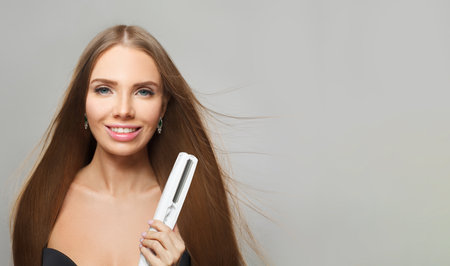 Nice fashion model with brown shiny straight haistyle portrait. Cute woman with straightening iron in her hand on gray studio backgroundの写真素材
