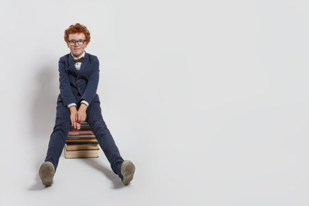Clever redhead child school kid sitting on stack of books against white studio wall background with copy spaceの写真素材