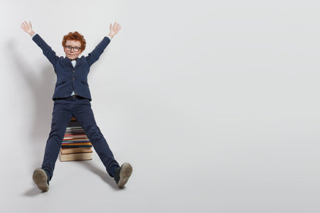Happy redhead child school kid sitting on stack of books and having fun against white studio wall background with copy spaceの写真素材