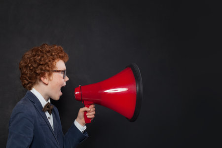 Funny cute little child boy student in school uniform speaking through megaphone against on black background. Idea, success and education conceptの写真素材