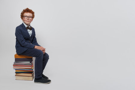 Intelligent child schoolboy sitting on stack of books and looking at camera against white studio wall background with copy spaceの写真素材