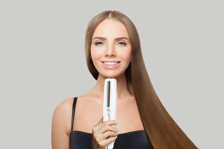 Cheerful young female model woman with long brown smooth hairstyle holding straight iron. Hairdresser, beauty care, hair straightening, cosmetic and stylingの写真素材
