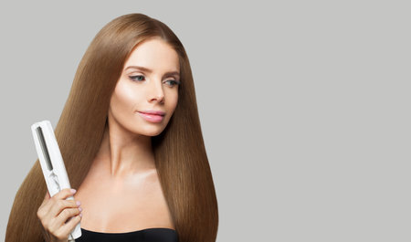 Gorgeous brunette woman holding straight hair iron and straightening her long smooth shiny hair on white background. Haircare, hairstyle, hairdressing and hair styling conceptの写真素材