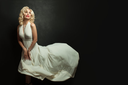 Glamorous woman with blonde hair in white silky dress on black background. Beautiful blond lady with make-up and wavy hairstyleの写真素材