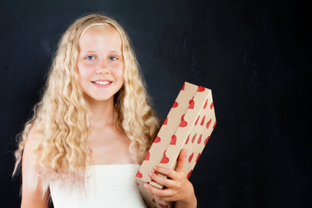 Attractive young girl holding gift box with red heart. Blond child with long healthy curly hair on black backgroundの写真素材