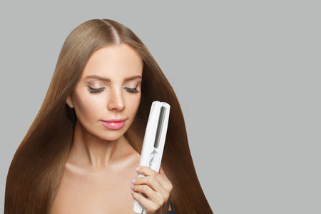 Brunette woman holding straight hair iron and straightening her long smooth shiny hair on white background. Haircare, hairstyle, hairdressing and hair styling conceptの写真素材
