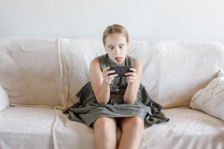 Funny young girl using the phone and sitting on sofa at cozy home. Child and smartphone. Technology,...