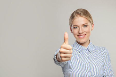Successful business woman looking at camera, smiling and holding thumb up on gray background with copy spaceの写真素材