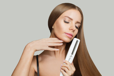 Glorious smiling woman with long straight healthy hairstyle using straightening iron on white background. Beauty, hair care, hair treatment and cosmetic conceptの写真素材