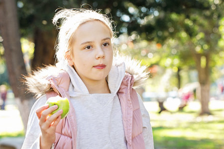 Cheerful young girl eating green apple outdoors, portrait. Child 11 years oldの写真素材